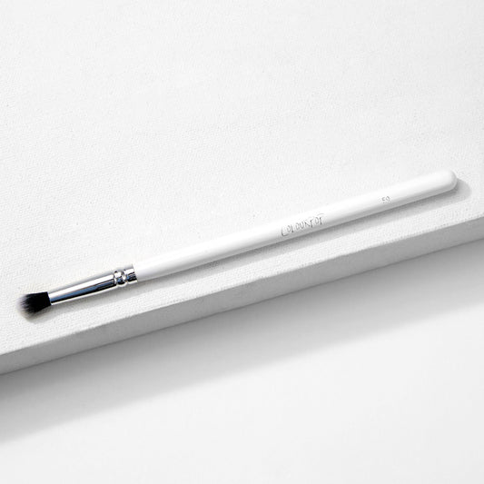 ColourPop Small Tapered Brush resting diagonally on a white textured platform, featuring a slim white handle, chrome ferrule, and small tapered black-and-white bristles for precise eyeshadow blending and detail work.